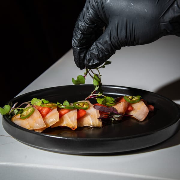 A gloved hand garnishes a black plate of salmon sashimi with microgreens, jalapeños, and red sauce.