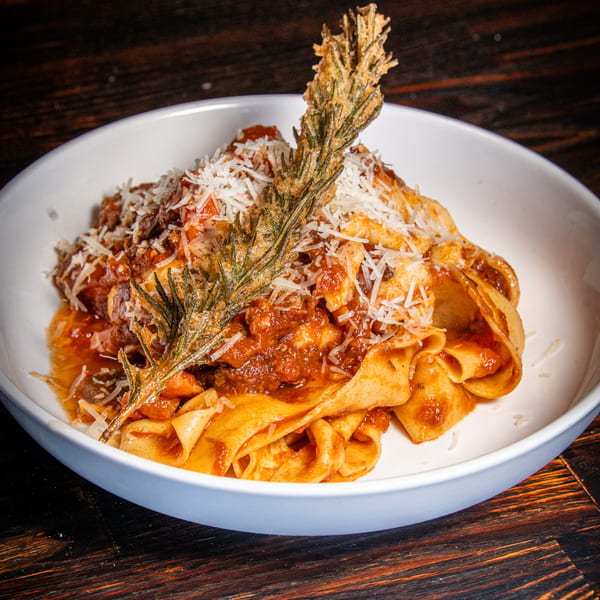 Pappardelle pasta with rich red sauce, grated cheese, and a fried rosemary sprig in a white bowl.
