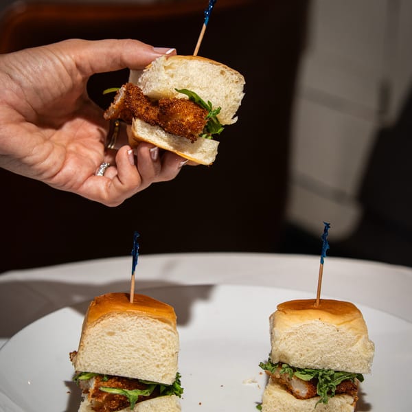 A hand holds a mini fried chicken slider above two more on a white plate.
