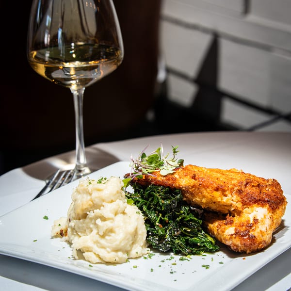Crispy fried chicken, mashed potatoes, and sautéed greens on a white plate with a glass of white wine.