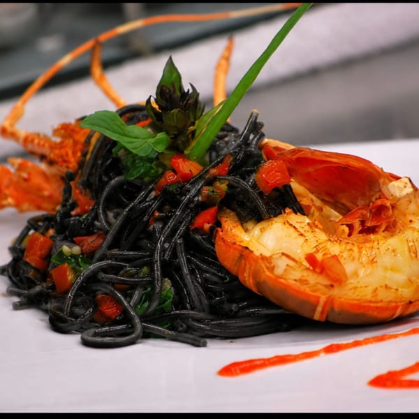 Black squid ink pasta with diced tomatoes, basil, and a grilled lobster tail on a white plate.