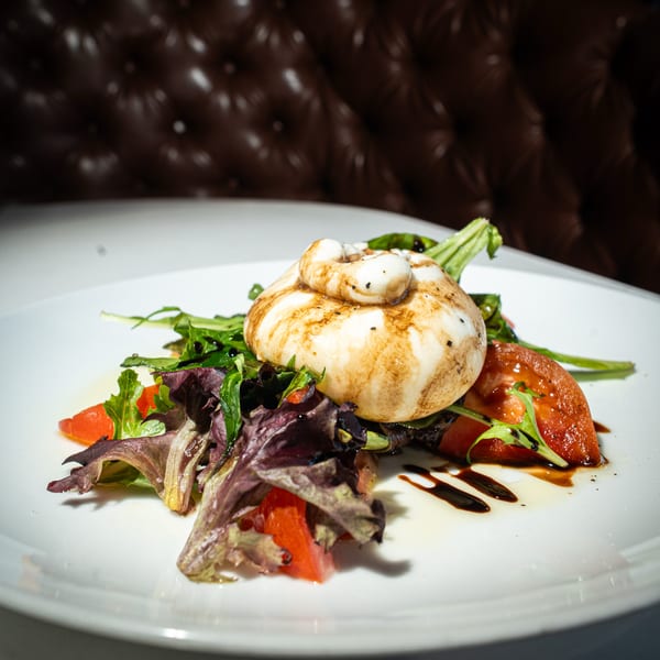 Burrata cheese with balsamic glaze, mixed greens, and sliced tomatoes on a white plate.