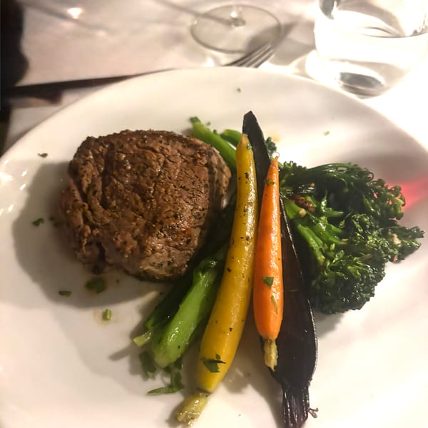 A steak entree with broccolini and colorful carrots on a white plate at a restaurant.