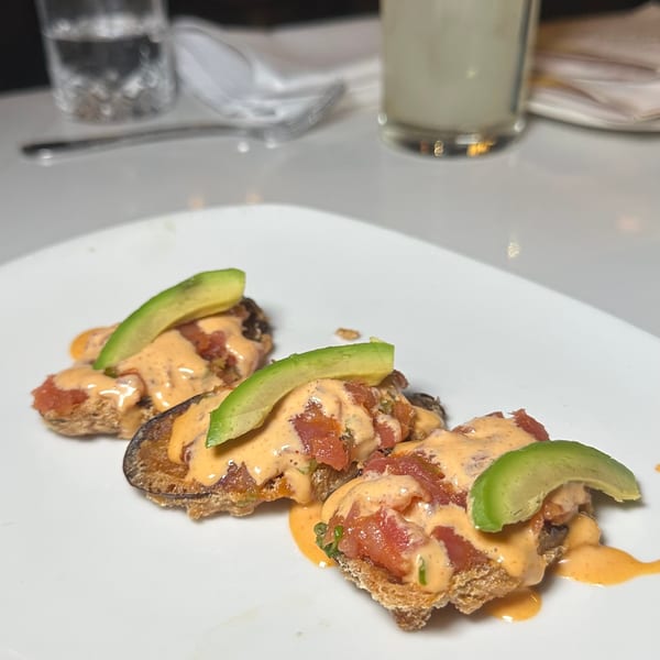 Three appetizers with diced tomatoes, creamy sauce, and avocado slices on a white plate, with drinks in the background.
